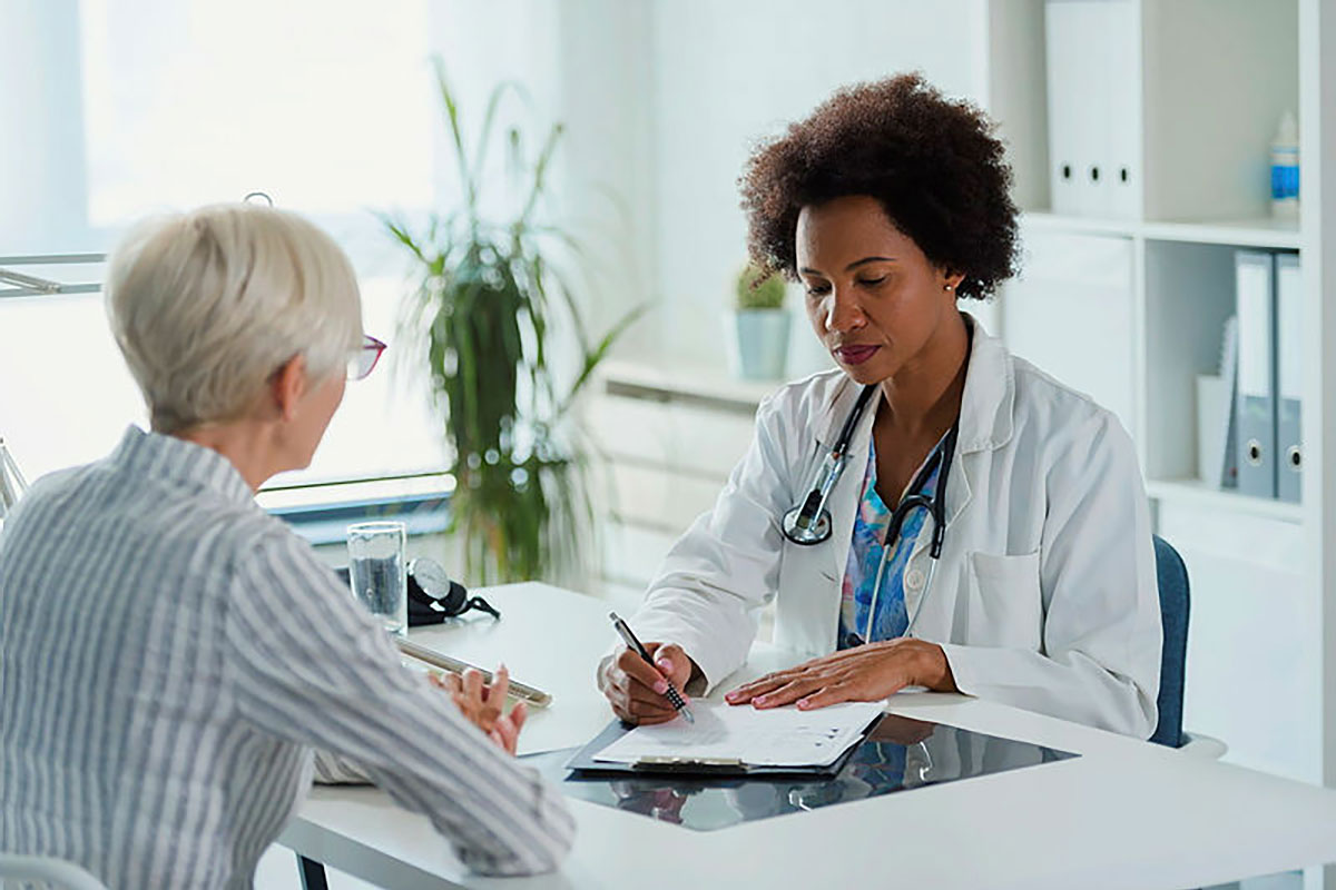 A female doctor sits at her desk and chats to an elderly female patient.