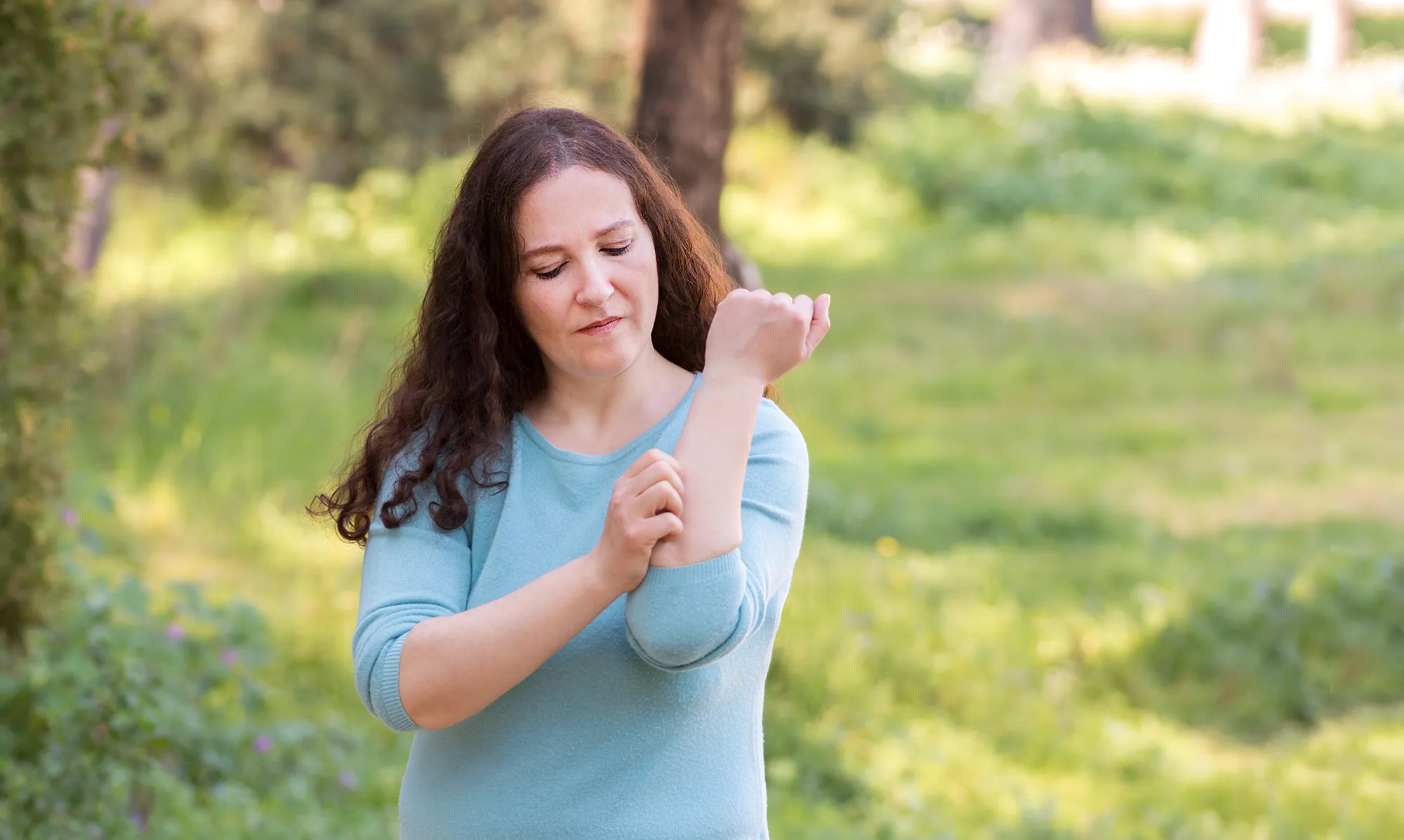 Person standing outdoors in a grassy, tree-filled area, using one hand to scratch the forearm of the other arm, wearing a light blue long-sleeve shirt.