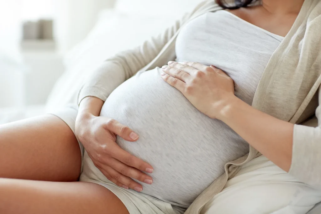 A close-up view of a pregnant person sitting on a bed, wearing a light-colored top and cardigan, with both hands resting on their prominently pregnant abdomen.