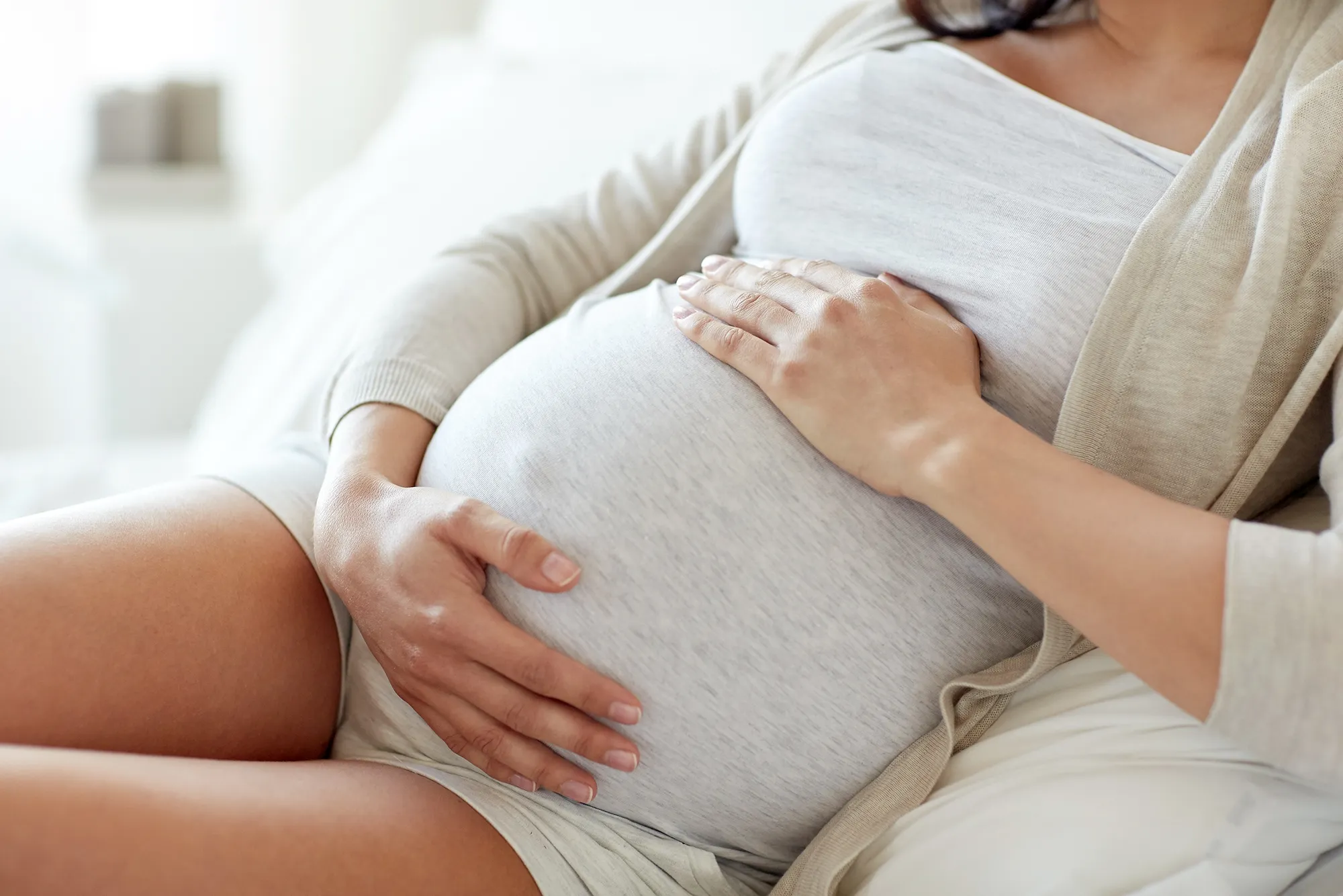 A close-up view of a pregnant person sitting on a bed, wearing a light-colored top and cardigan, with both hands resting on their prominently pregnant abdomen.