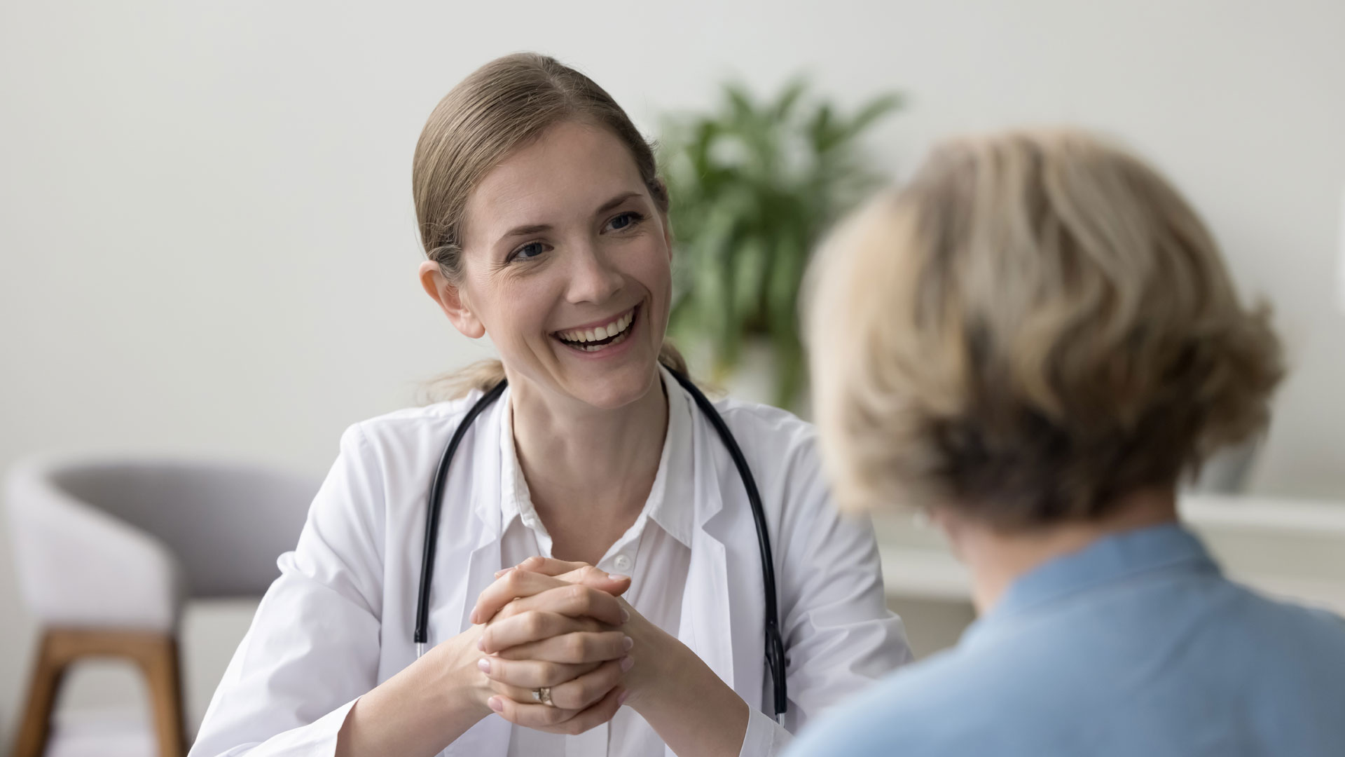 Healthcare professional in a white coat with a stethoscope around the neck sitting across from a patient during a consultation in a bright, modern office setting.