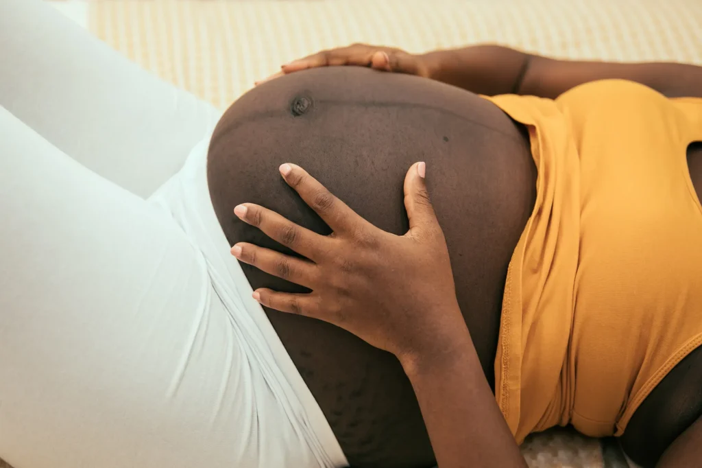 Close-up of a pregnant abdomen with both hands resting on and gently holding the belly, with a light-colored bed or cushion in the background and a sleeveless top visible.